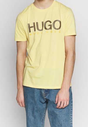 Mand iført en gul kortærmet T-shirt med "HUGO BOSS" logo i store bogstaver og blå denim jeans.