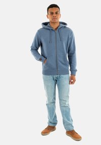 Blå zip-up hættejakke med snoretræk på hætten, forlommer og ribbede manchetter, parret med lyseblå jeans og brune sko.