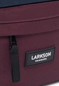 Bolsa de tela color burdeos con parte superior azul marino, que cuenta con un cierre de cremallera, etiqueta negra con "LARKSON HAMBURG" y superficie texturizada.