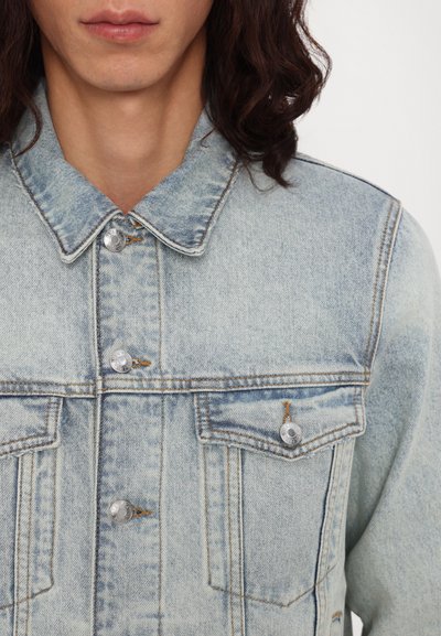 Light blue denim jacket with a collar, front button closure, and two chest pockets. Features silver buttons and faded texture.