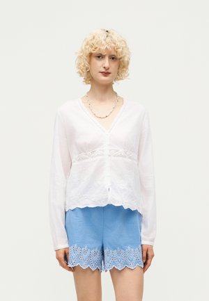 Osoba s kovrčavom plavom kosom koja nosi bijelu bluzu dugih rukava s detaljima čipke i plave šivane kratke hlače, stojeći ispred jednobojne pozadine.