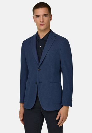Giovane uomo che indossa un blazer blu navy sopra una polo nera e pantaloni neri, in piedi davanti a uno sfondo chiaro e uniforme.