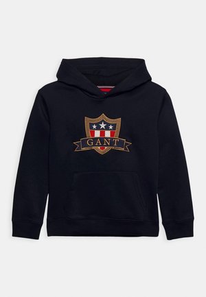 Navy blue hoodie made of cotton, featuring an embroidered shield design with stars and a banner reading "GANT" on the front.