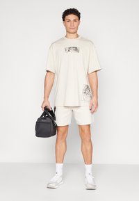 T-shirt gráfica bege com dois desenhos impressos, combinada com calções brancos. Uma bolsa preta segurada numa das mãos e ténis claros com meias brancas.