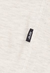 Lichtgrijs ribbeltricot met een klein zwart label waarop "J&Joy" staat, genaaid in de zijnaad. De textuur lijkt zacht en warm.