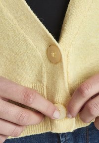 Gul stickad cardigan med V-ringning och två stora, krämfärgade knappar fram. Texturen är mjuk med en ribbad nederkant.