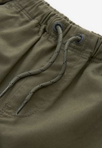 Shorts vert olive en tissu texturé, dotés d'une taille élastique avec des cordons noirs et des embouts argentés pour le réglage.