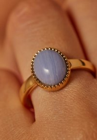 Gouden ring met een gepolijste, ovale lichtpaarse steen. De steen is omringd door een kralenachtige gouden setting, rustend op een gladde band.