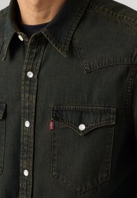 Camicia in denim verde scuro con colletto a punta, bottoni a pressione e due tasche sul petto con bottoni bianchi. Etichetta del logo Levi's sul lato.
