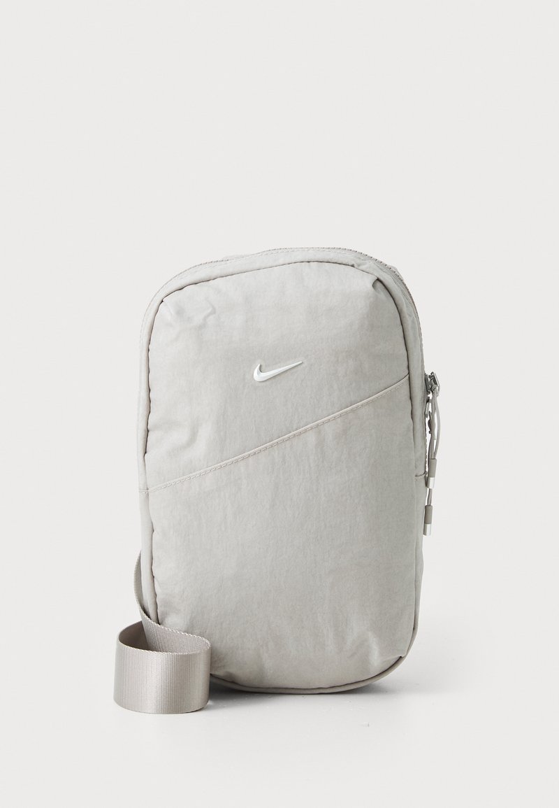 Pelēks nailona plecu soma ar ovālu taisnstūra formu, ar rāvējslēdzēja aizdari, diagonālu šuvju un baltu Nike logo priekšpusē.
