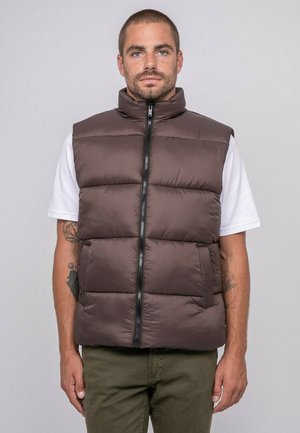 Homme aux cheveux courts et tatouages, portant une veste matelassée marron sur une chemise blanche à manches courtes et un pantalon vert, debout devant un fond gris.