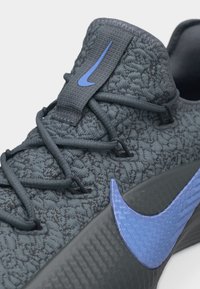 Κοντινό πλάνο αθλητικού παπουτσιού σε σκούρο γκρι με υφή, με μπλε λογότυπο Nike στη σόλα και τη γλώσσα, που διαθέτει μοναδικό σύστημα κορδονιών και πλεκτό επάνω μέρος.