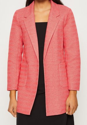 Donna che indossa un blazer rosso a spina di pesce sopra un vestito nero, mostrando la trama del blazer, le rever, le maniche lunghe e le tasche frontali.