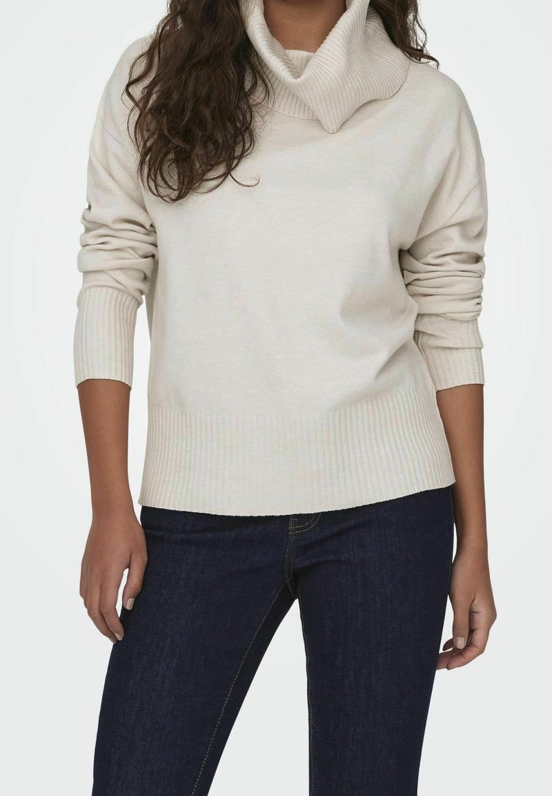 Femme portant un pull à col roulé crème avec des poignets et un bas côtelés, associé à un jean bleu foncé sur un fond blanc uni.