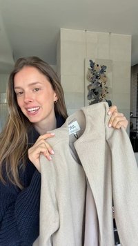 Jeune femme aux longs cheveux tenant un manteau beige, souriant à l’intérieur avec une œuvre d'art abstraite sur le mur derrière elle.