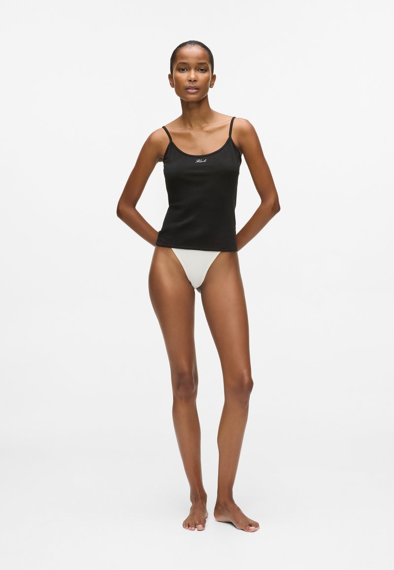 Melnā piegulošā korsetē ar šaurām siksnām, priekšpusē ar nelielu baltu logo, apvienota ar gaišas krāsas peldbiksēm.