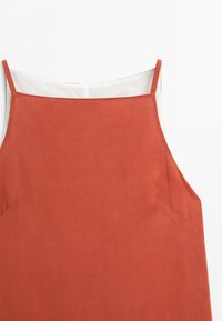 Mouwloze jurk in roestkleur met dunne bandjes en een vierkante halslijn over een witte voering die bovenaan zichtbaar is.