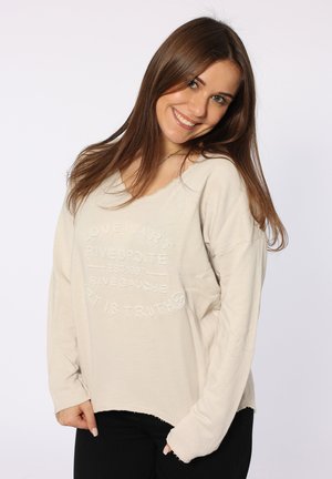 Helles beiges Langarmshirt mit lockerem Schnitt, das mit geprägtem Text und fransigem Saum versehen ist, kombiniert mit schwarzen Hosen.