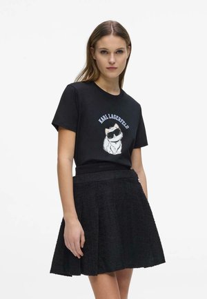 Noor naine kannab musta Karl Lagerfeld T-särki, millel on kassi graafika, ja musta tekstuurse A-lõikega seelikut, taustaks ühevärviline valge taust.