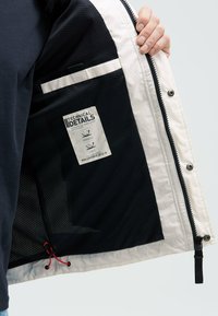 Interno della giacca bianca e nera, con fodera in rete, una tasca con zip e un'etichetta con dettagli tecnici. Coulisse all'orlo.