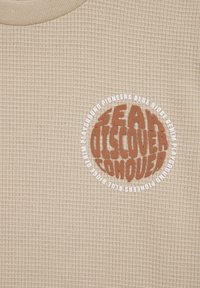 Chemise beige à texture gaufrée avec un logo circulaire portant du texte marron et blanc : « SEAN DÉCOUVRIR CONQUÉRIR PIONNIERS DE BLUE RIDGE ». 