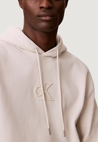 Felpa con cappuccio beige chiaro con logo "CK" in rilievo, coulisse e tessuto morbido. Presenta una vestibilità comoda e un design minimalista.
