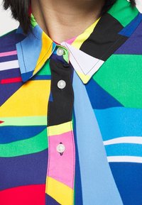 Kolorowa koszula z kołnierzykiem, zdobiona wzorami geometrycznymi. Wykonana z gładkiego materiału, z guzikami z przodu. Dominujące kolory to niebieski, zielony i żółty.