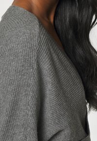 Pull en maille côtelée gris avec un décolleté en V profond et des manches amples, présentant une texture douce et une silhouette drapée.