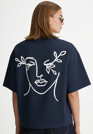 Donna con capelli fino alle spalle che indossa una camicia blu scuro con un motivo astratto bianco di volto e foglia sulla schiena, vista di profilo.