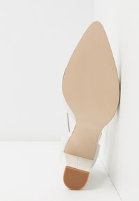 Semelle beige pointue d'une chaussure blanche à talon haut avec un petit talon marron texturé appuyé contre un coin blanc.