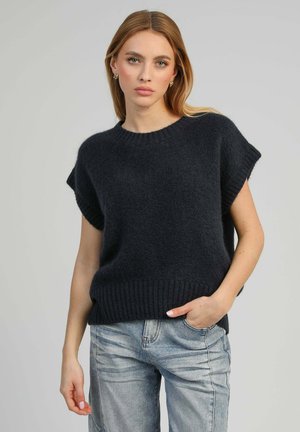 Jeune femme aux cheveux longs et blonds portant un pull sans manches noir en maille ample et un jean bleu clair, posant avec une main dans la poche.