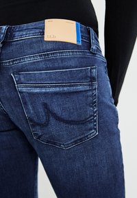 Blaue Denim-Jeans mit glatter Textur, ausgestattet mit einem braunen Lederetikett, zwei Gesäßtaschen und Kontrastnähten.