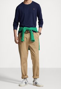 Námořnická košile s dlouhým rukávem, zelený svetr uvázaný v pase, béžové chinos kalhoty, bílé tenisky s černými pruhy, hodinky na zápěstí, volný střih.