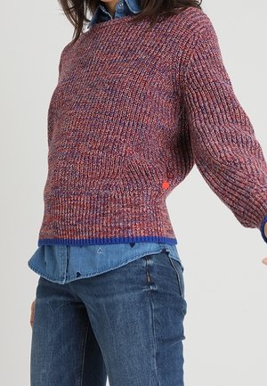 Vrouw draagt een meerkleurige gebreide trui over een blauw overhemd, gecombineerd met donkerblauwe jeans, tegen een effen achtergrond.