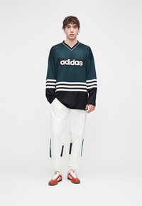 Fekete és türkiz hosszú ujjú mez fehér csíkokkal és „adidas” logóval, fehér nadrággal, amely kiemelő panelekkel és piros tornacipőkkel van párosítva.