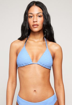 Frau mit langen dunklen Haaren, die ein strukturiertes hellblaues Bikini-Oberteil in Dreiecksform und passenden Bikini-Slip vor neutralem Hintergrund trägt.