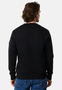 Svart sweatshirt med rund halsringning, texturerat tyg och en ficka på vänster ärm. Parat med blå denimjeans, visar en avslappnad bakvy.