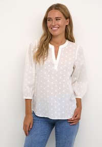 Blusa blanca con un patrón de lunares texturizados, tela ligera, escote redondo y mangas abullonadas de tres cuartos. Combinada con jeans azules.