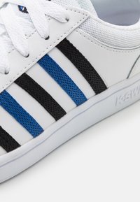 Białe sneakersy z niebieskimi i czarnymi teksturowanymi paskami, białymi sznurówkami oraz białą podeszwą z wytłoczoną nazwą "K-SW".