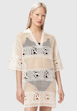 Person med langt blondt hår iført en hvid hæklekjole med "SAS"-mønster over sorte undertøj, stående foran en ensfarvet baggrund.