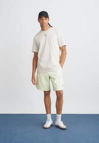 Tricou bej deschis cu un logo mic, șorturi verde mentă cu detalii de cusătură, adidași albi și o șapcă neagră. Ținută simplă, casual, sport.