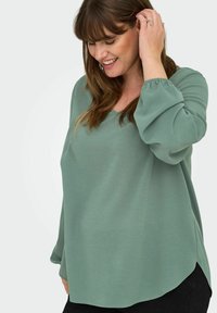 Zaļa garo piedakļu blūze ar V-veida kaklu, izgatavota no viegla auduma. Tai ir spuldžu piedurknes un brīva piegriezuma siluets ar nedaudz izliekta apakšmalu.