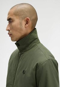 Giacca verde oliva con colletto alto, dotata di chiusura a zip e un piccolo logo ricamato sul petto. Tessuto liscio e vestibilità su misura.