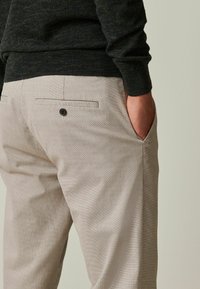 Pantalones texturizados de color beige claro con un patrón sutil, que cuentan con un bolsillo trasero y un botón. Suéter verde oscuro atado en la cintura.