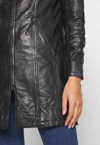 Schwarzer Ledermantel mit strukturiertem Finish, vertikalen Nahtdetails und zwei seitlichen Reißverschlusstaschen. Lange Ärmel und mittellange Schnittführung.