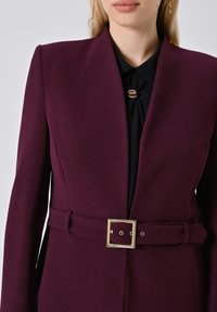Burgunderfarbener maßgeschneiderter Blazer mit tiefem V-Ausschnitt, einem Stoffgürtel mit goldener quadratischer Schnalle und langen Ärmeln über einer schwarzen Bluse.