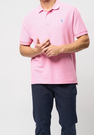 Man wearing a pink short-sleeve polo shirt with a small blue logo and dark navy pants, hands clasped in front.
