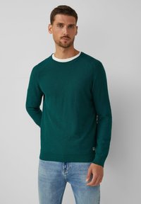 Groene gebreide trui met lange mouwen, ronde hals en een contrasterende witte binnenkraag. Strak model, gecombineerd met lichtblauwe jeans.