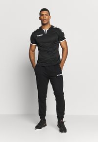 Sort sports skjorte med hvide detaljer og logo, parret med sorte sweatpants med logo, båret med sorte træningssko.