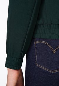 Mørkegrøn bluse med tekstureret stof, pufærmer og elastiske manchetter, parret med mørkeblå jeans med gylden syning på lommen.